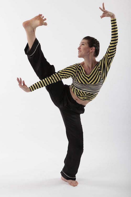 The dancer's left leg is raised high in the air with her toes flexed. Her eyes look towards the ceiling.